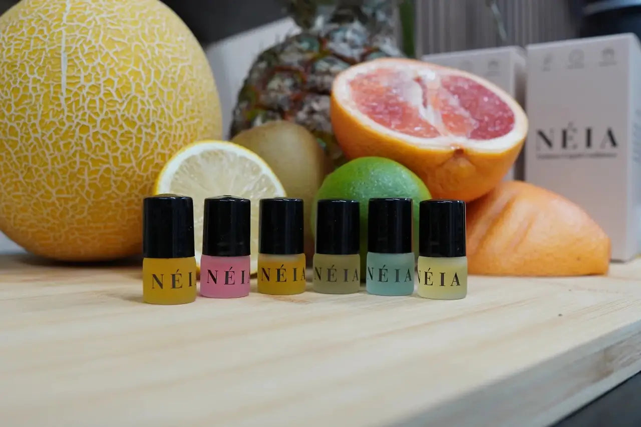 The six oils on a wooden counter in front of a melon, a pineapple and a halved grapefruit.