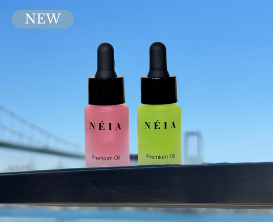 Two NÉIA premium oil dropper bottles, one pink and one green, on a reflective surface with a blue sky background.