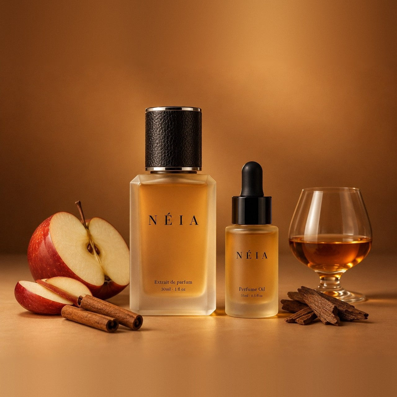 The Apple Cognac layering kit: the 30ml NÉIA Extrait de Parfum beside the 15ml Perfume Oil, with a halved red apple, cinnamon sticks, oak chips and a glass of cognac.